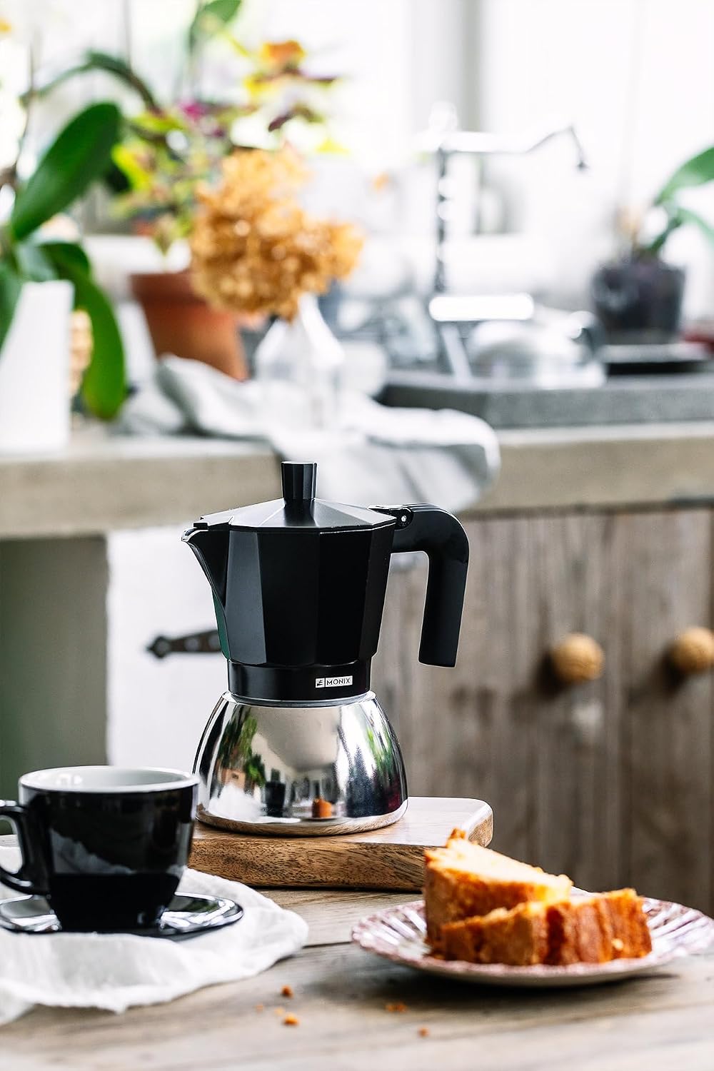 Cafetera italiana Monix negra en una mesa de cocina junto a una taza de café y un bizcocho